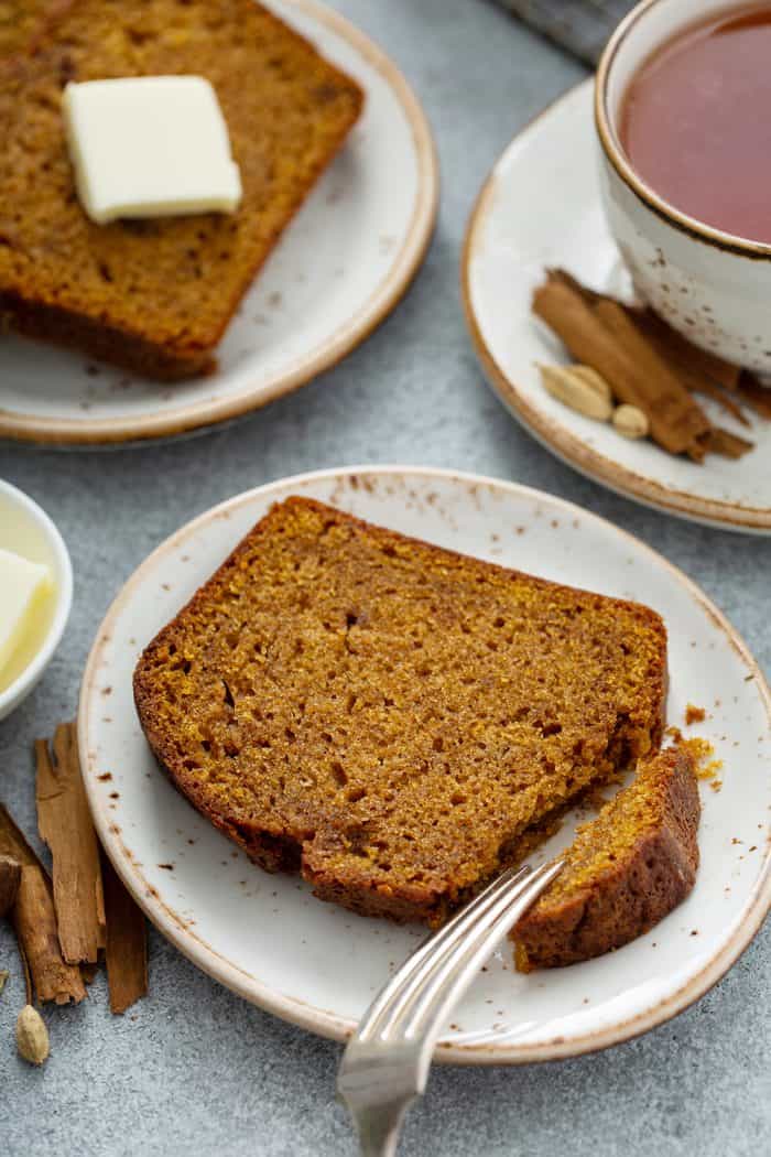 Fork cutting into a slice of butternut squash bread on a white plate, with a cup of chai and another slice of bread in the background