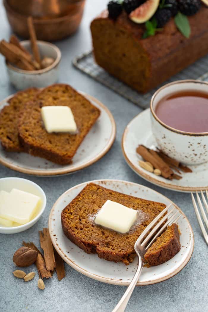 Two plates with butter-topped slices of butternut squash bread with a cup of chai and a loaf of bread in the background