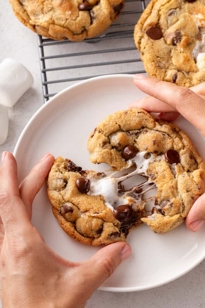Hand pulling apart a giant s'mores cookie on a white plate to show the gooey chocolate and marshmallow.