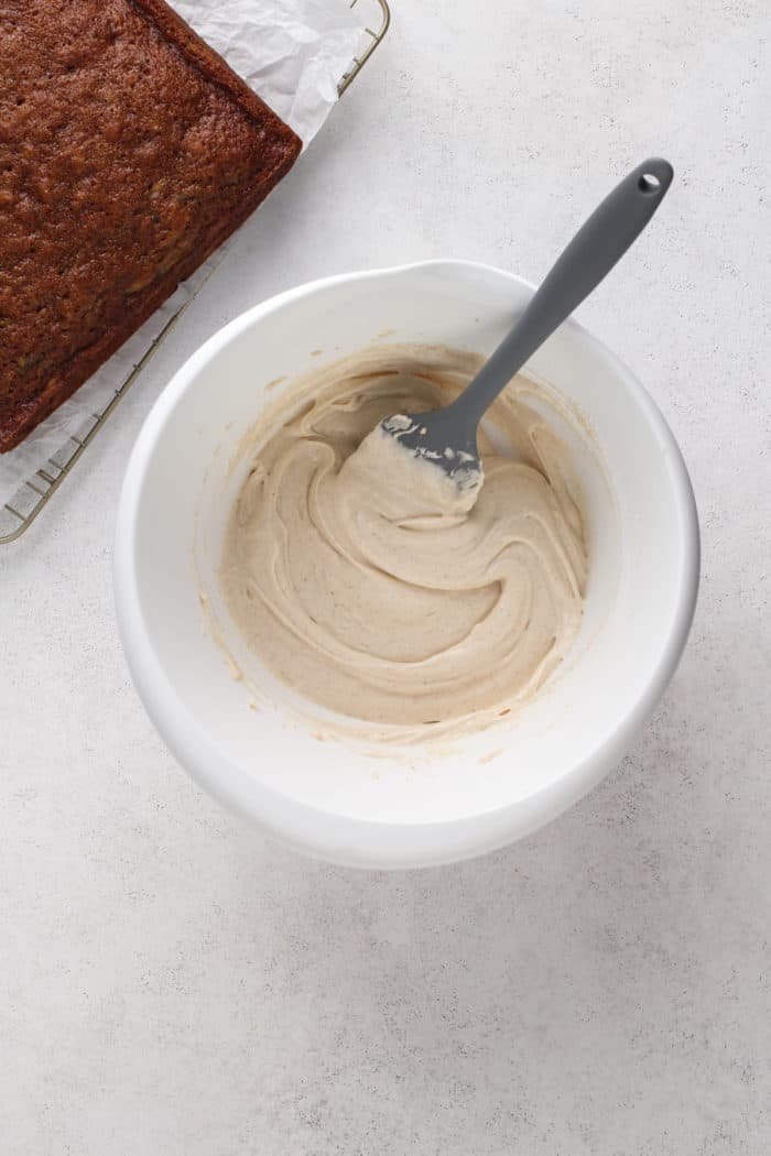 Baked and cooled zucchini cake on a cooling rack next to a bowl of maple cream cheese frosting.