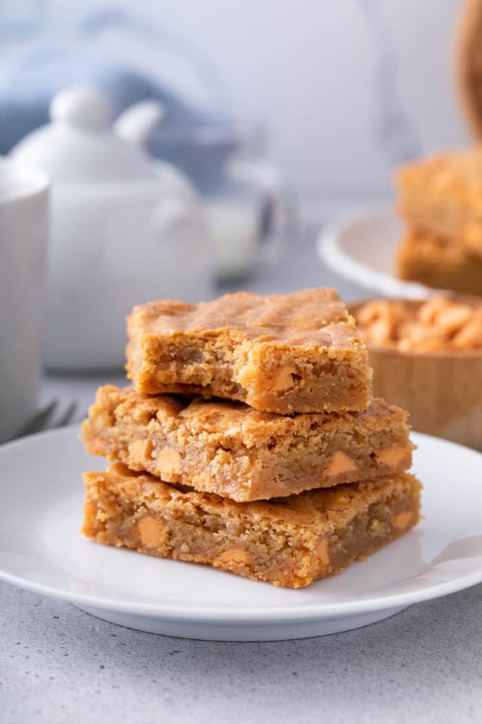 Three brown butter blondies stacked on a white plate with a bite taken out of the top blondie.