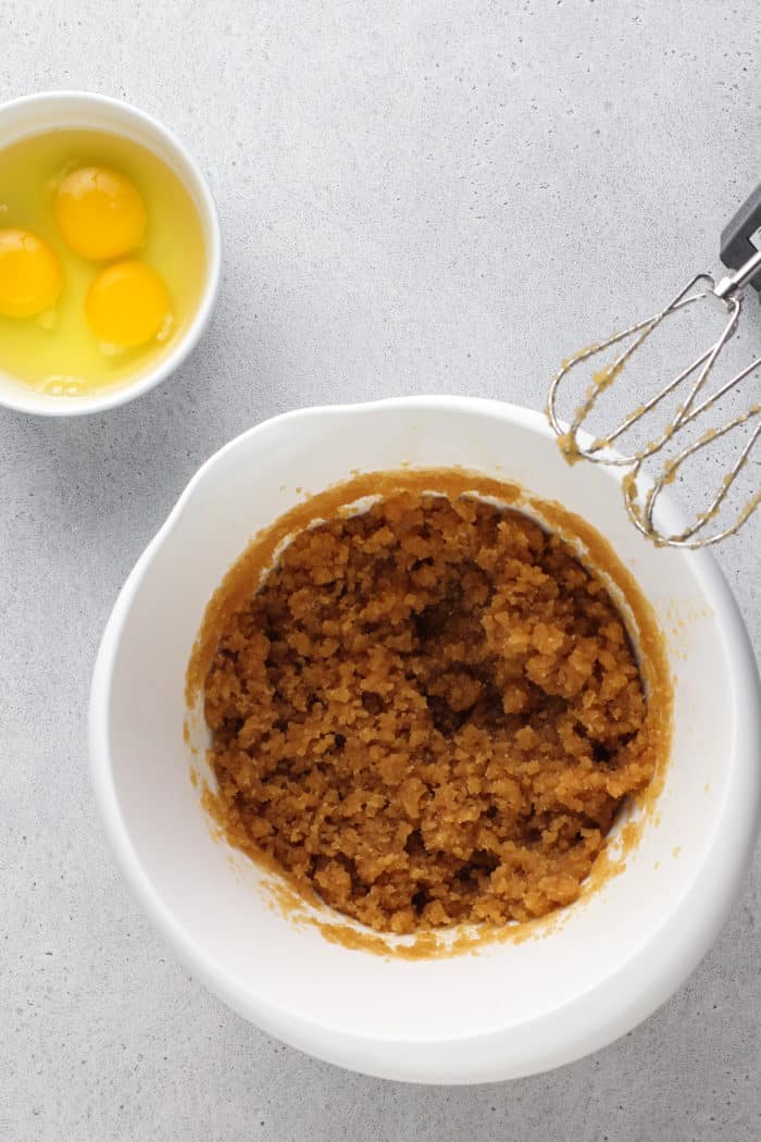 Brown butter and sugar mixed together in a white mixing bowl set next to a bowl of eggs.