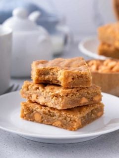 Three brown butter blondies stacked on a white plate with a bite taken out of the top blondie.