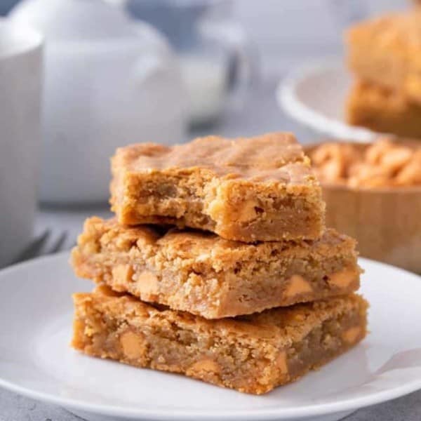 Three brown butter blondies stacked on a white plate with a bite taken out of the top blondie.