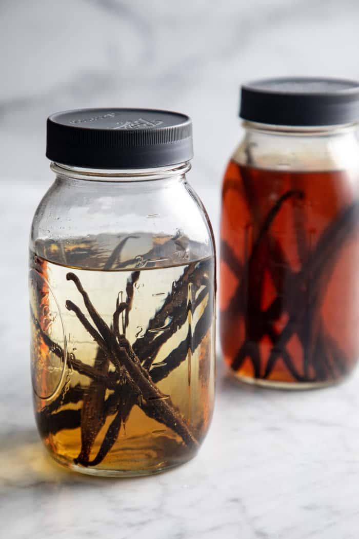 Two large mason jars of vanilla extract. One jar is still infusing and is light in color, while the second one is done infusing and is dark in color.