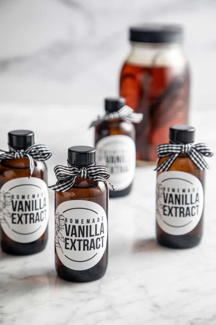 Four small, labeled bottles of homemade vanilla extract with a large jar of the extract visible in the background.