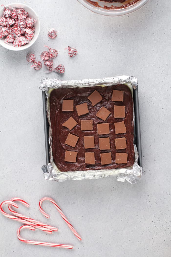 Squares of a milk chocolate bar on top of a layer of peppermint brownie batter in a foil-lined square pan.