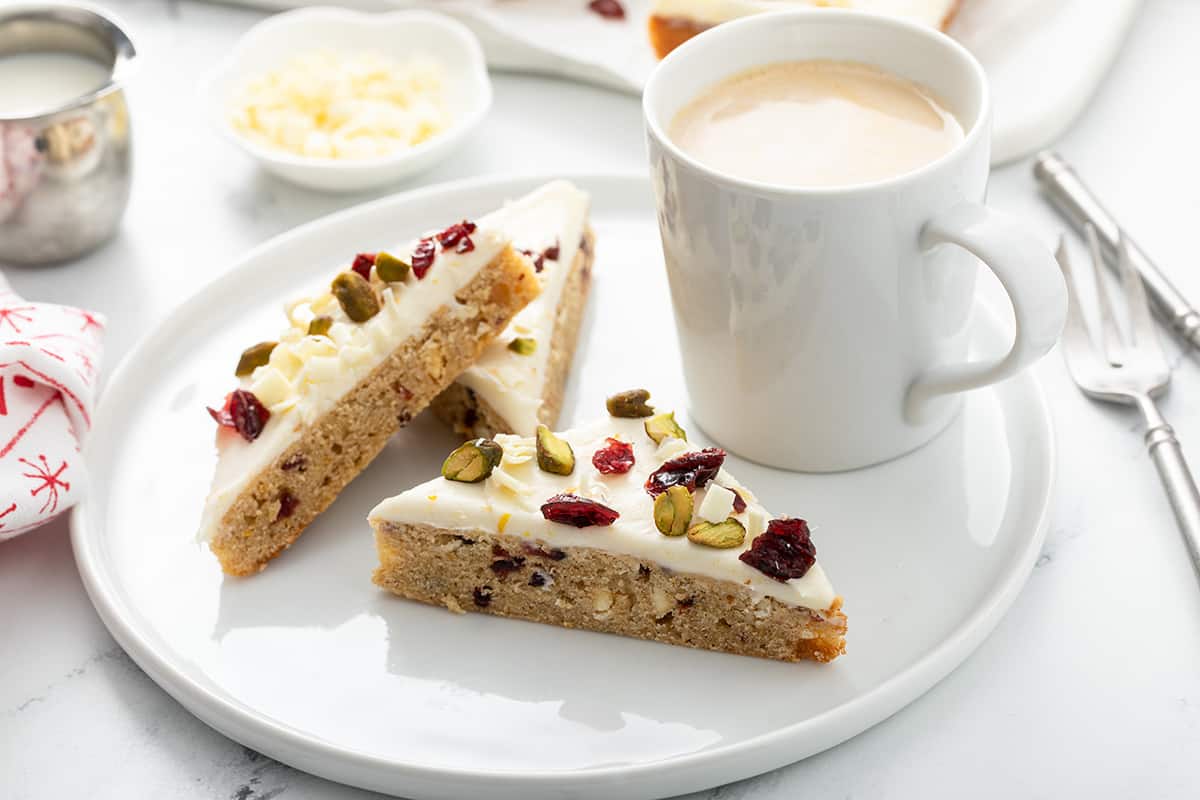 Three cranberry bliss bars set next to a cup of coffee on a white plate