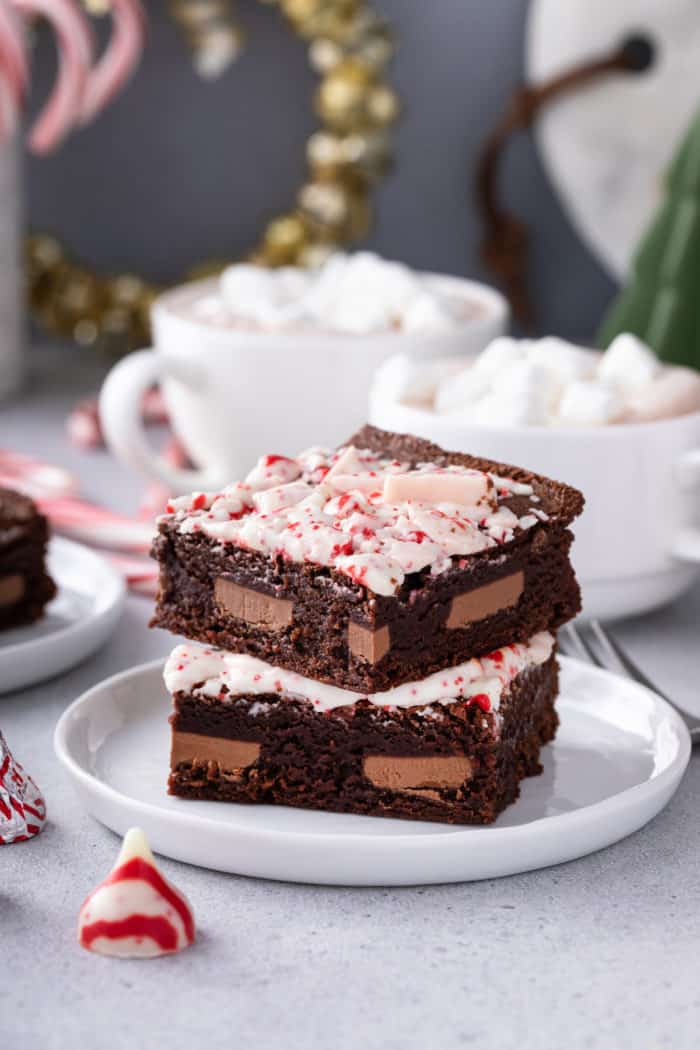 Two stacked peppermint brownies on a white plate, with cups of hot cocoa in the background.