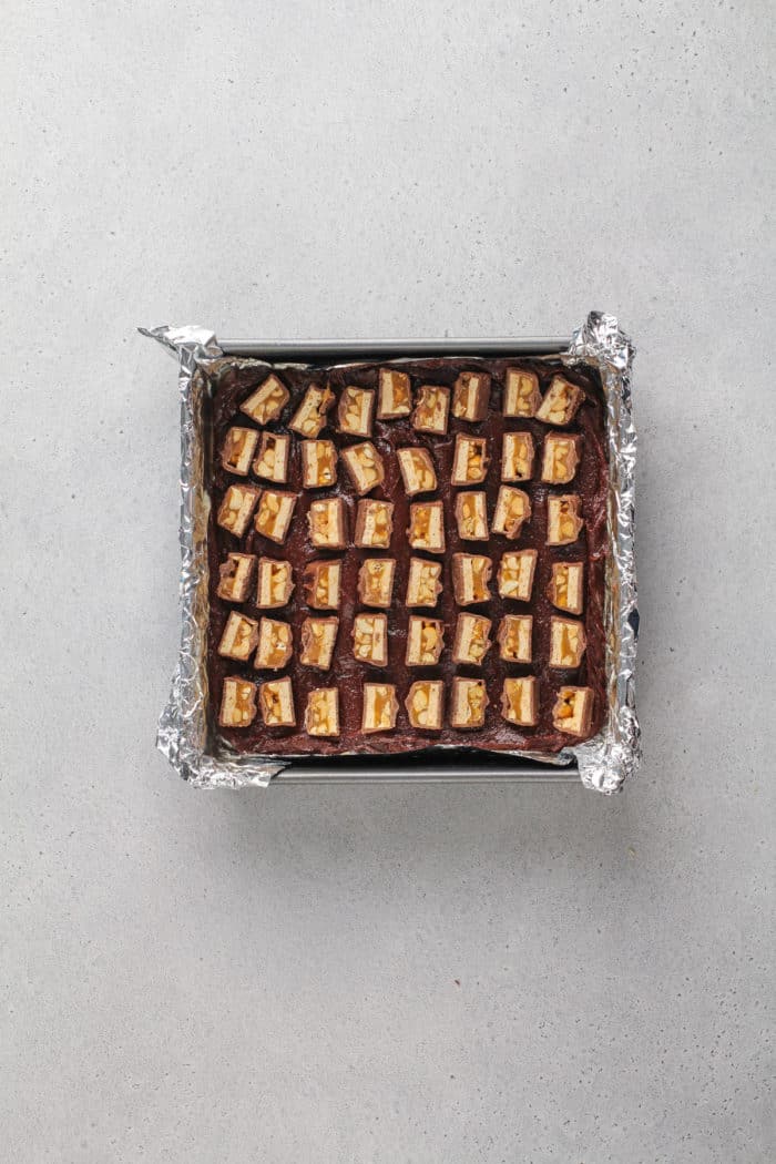 Pieces of snickers bars arranged on top of brownie batter in a foil-lined pan.