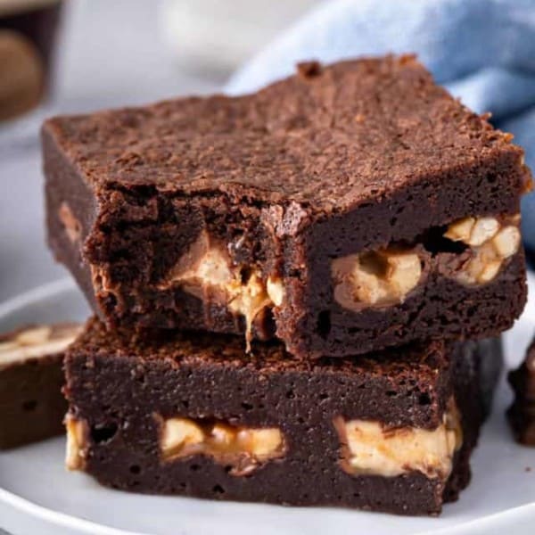 Two snickers brownies on a white plate, with a bite taken out of the corner of the top brownie.