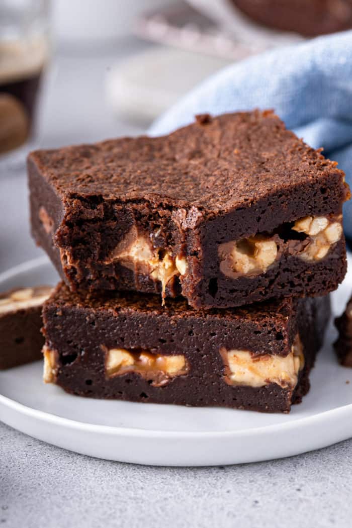 Two snickers brownies on a white plate, with a bite taken out of the corner of the top brownie.