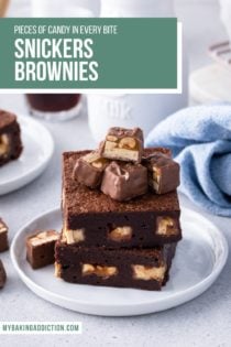 Two snickers brownies stacked on a white plate, topped with pieces of snickers bars. Text overlay includes recipe name.