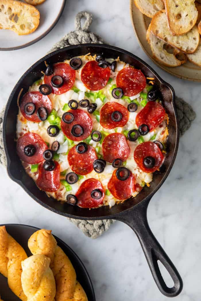 Baked pizza dip in a cast iron skillet, surrounded by bowls of crostini and breadsticks.