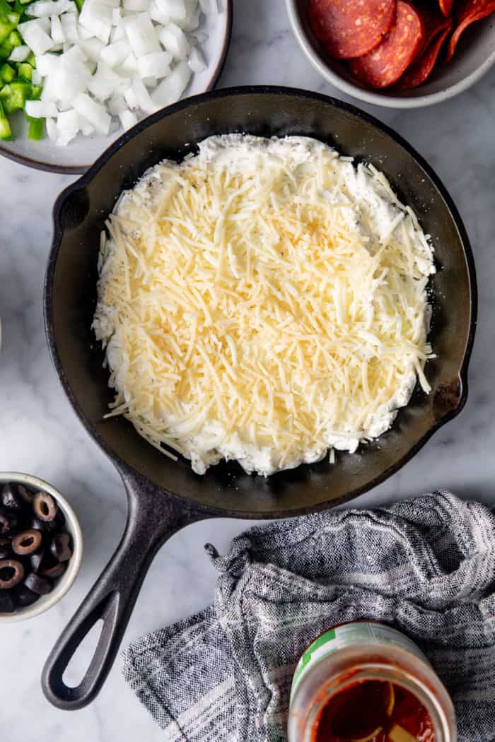 Herbed cream cheese topped with shredded cheese in a cast iron skillet on a marble countertop.