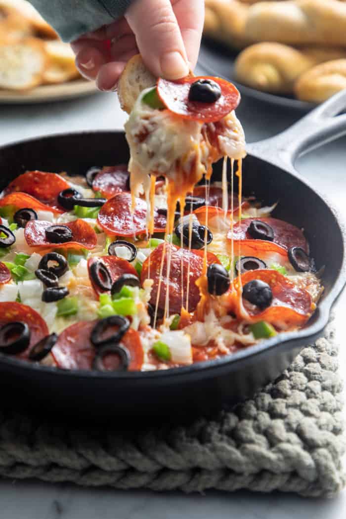 Hand dipping into a skillet of baked pizza dip with a crostini.