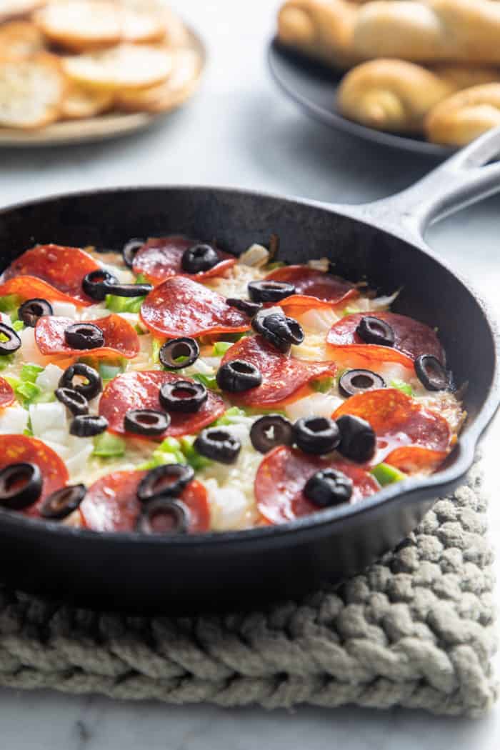 Pizza dip topped with pepperoni, olives, onions, and green peppers in a cast iron skillet on a beige trivet.