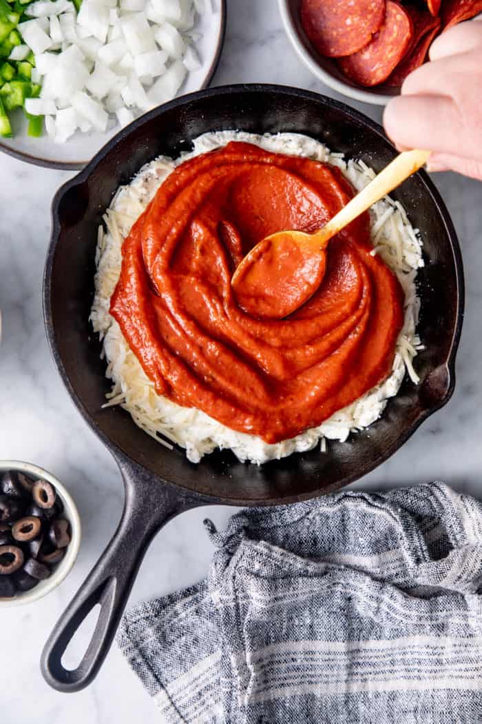 Pizza sauce being spread over shredded cheese and herb cream cheese in a cast iron skillet.