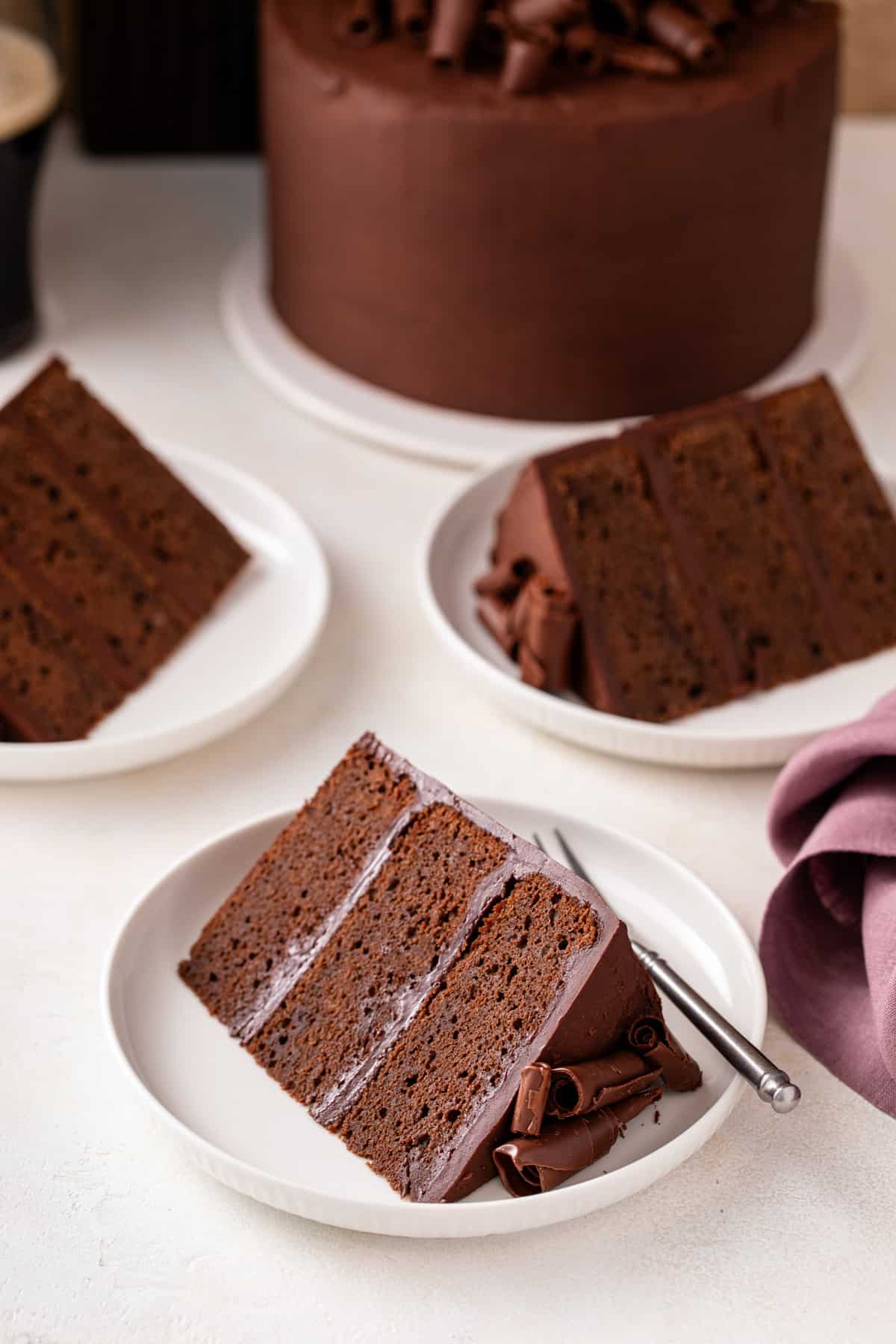 Three white plates, each holding a slice of chocolate guinness cake, with the rest of the cake in the background.