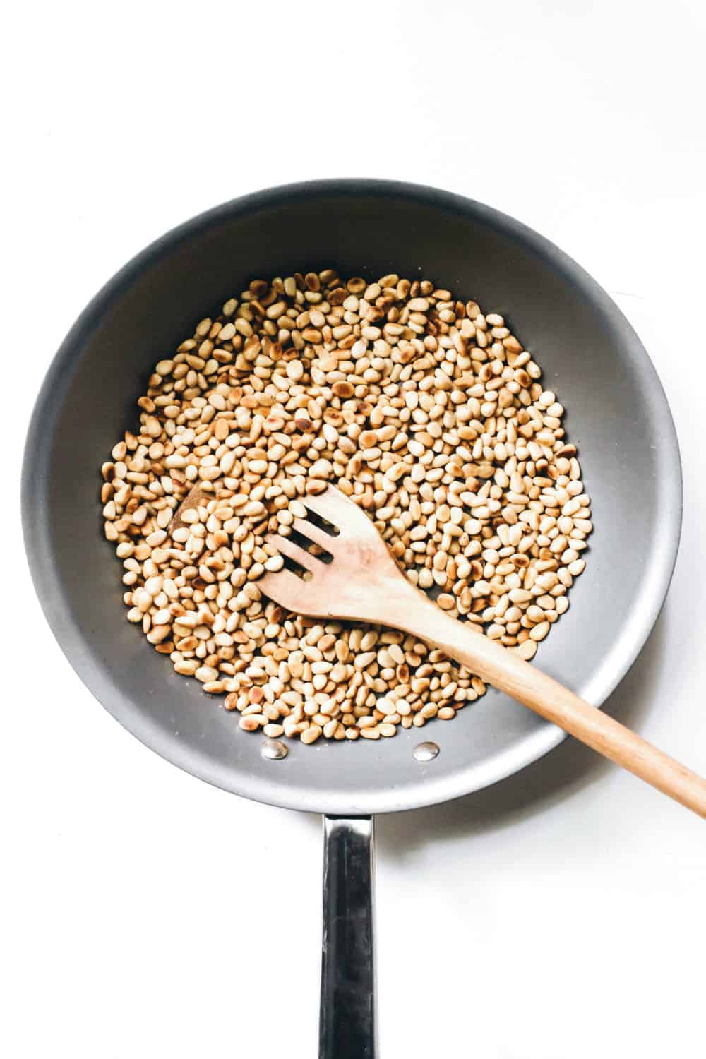 Toasted pine nuts in a skillet being stirred with a wooden spoon