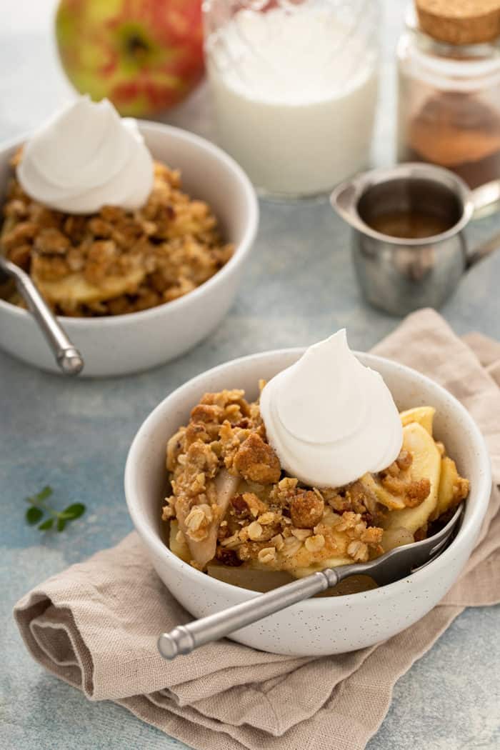 Two bowls of apple pear crisp, topped with whipped cream, on a countertop next to glasses of milk