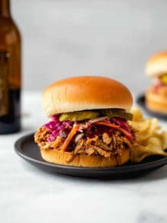 Pulled pork sandwich topped with slaw and pickles with a side of potato chips on a plate