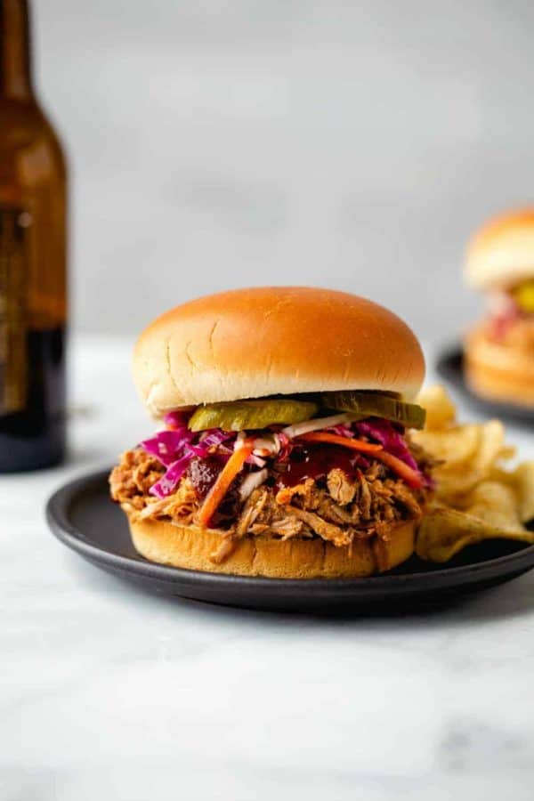 Pulled pork sandwich topped with slaw and pickles with a side of potato chips on a plate