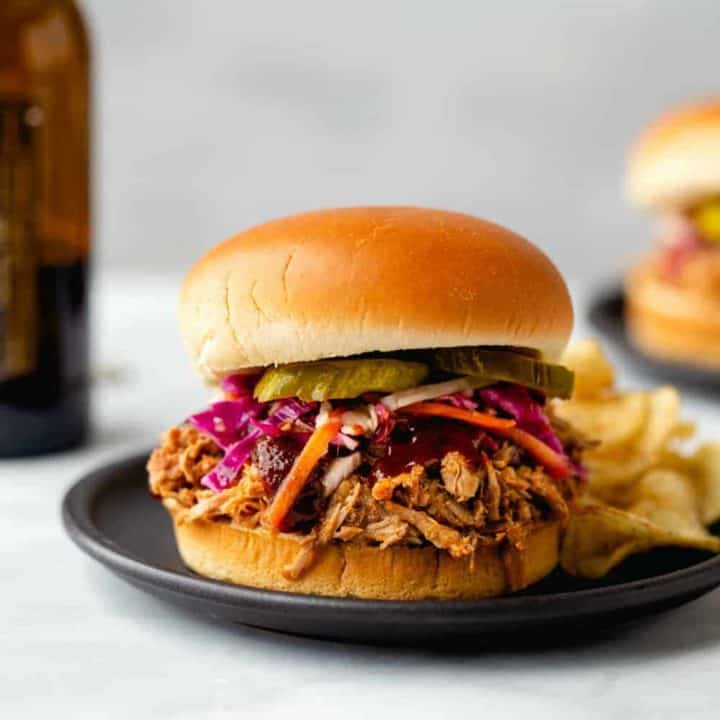 Pulled pork sandwich topped with slaw and pickles with a side of potato chips on a plate
