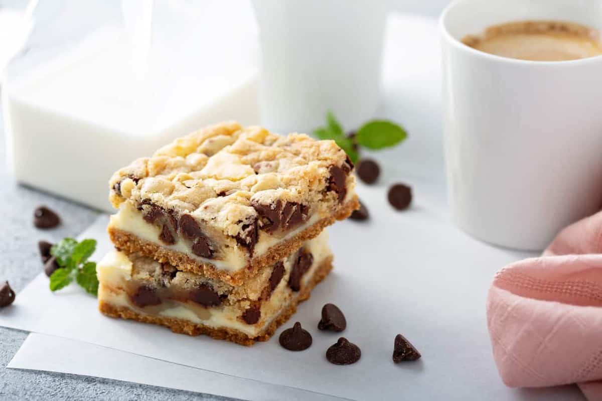 Two sliced cookie dough cheesecake bars on a piece of parchment paper next to a cup of coffee