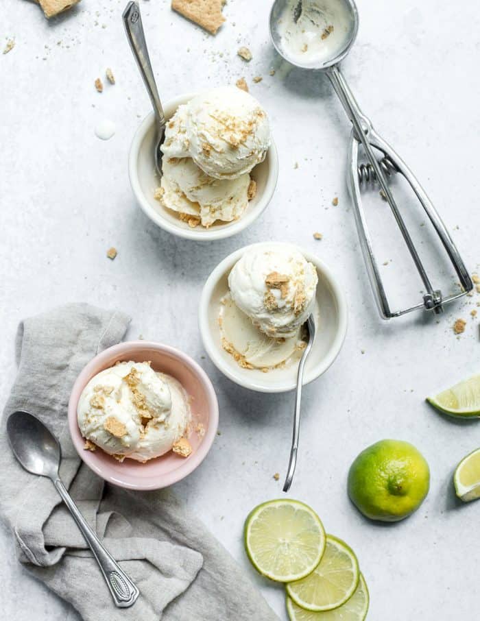 Three small bowls each filled with a scoop of key lime pie ice cream set on a marble counter next to an ice cream scoop and slices of limes