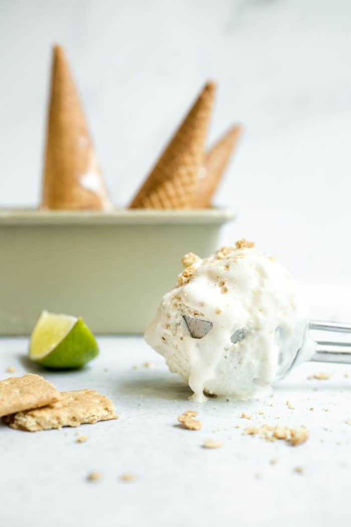 Ice cream scoop holding a melty scoop of key lime pie ice cream set on a marble counter with a container of sugar cones in the background