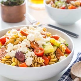Greek pasta salad topped with feta cheese in a bowl next to a plaid napkin with a wooden spoon