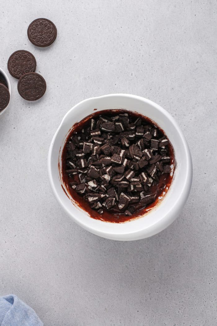 Chopped oreos being added to brownie batter in a white mixing bowl.