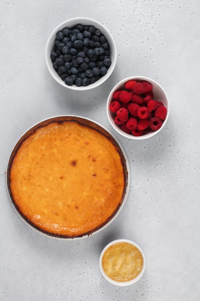 Baked ricotta cheesecake on a countertop next to bowls of raspberries and blueberries.