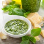 Small white bowl filled with basil pesto on a marble board surrounded by chunks of parmesan cheese and basil leaves