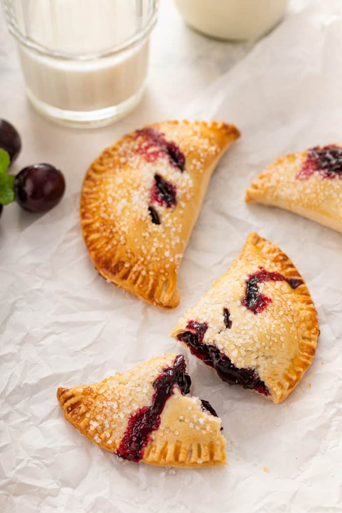 Two cherry hand pies on parchment paper next to a glass of milk. One of the hand pies is cut in half