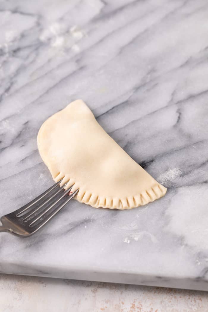 Fork crimping the edges of a hand pie sealed shut on a marble surface