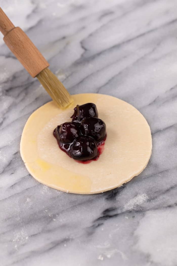 Egg wash being brushed on the edges of a circle of pie crust with cherry pie filling in the center