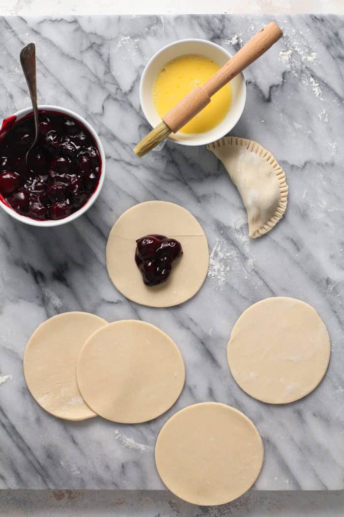 Circles of pie dough on a marble slab, with cherry pie filling in the center of one of the circles