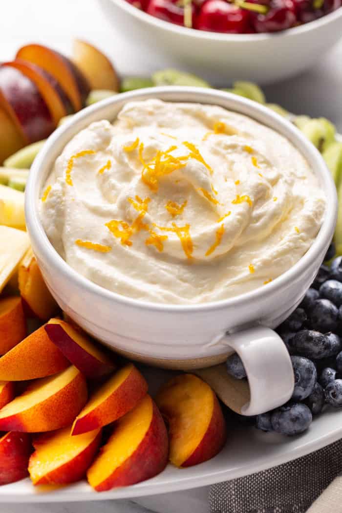 White bowl filled with orange fruit dip on a plate surrounded by cut fresh fruit