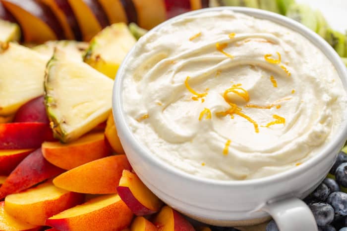 Close up of orange fruit dip in a white bowl, surrounded by cut fresh fruit