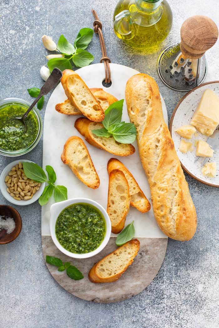 Overhead view of slices of baguette and a bowl of basil pesto on a marble serving board