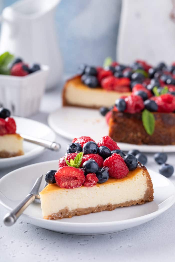 White plate with a slice of ricotta cheesecake that is topped with rows of raspberries and blueberries.