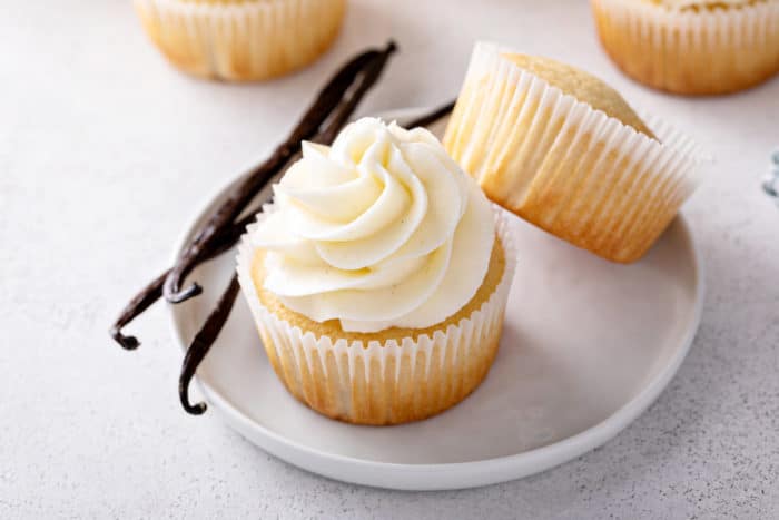 Two vanilla cupcakes set next to vanilla beans on a white plate. One of the cupcakes is frosted with vanilla frosting, the other is left unfrosted.