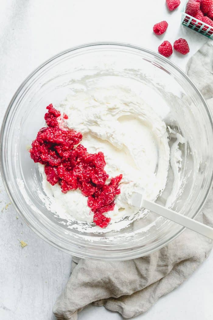 Spatula folding mashed raspberries into the filling for no-bake raspberry lemon cheesecake in a glass mixing bowl