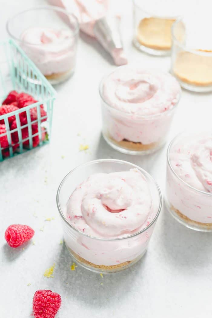 Three glass bowls of raspberry lemon cheesecake on a marble countertop next to a carton of fresh raspberries
