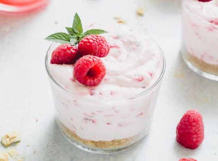 Close up of a glass dish of no-bake raspberry lemon cheesecake garnished with three raspberries and a sprig of mint