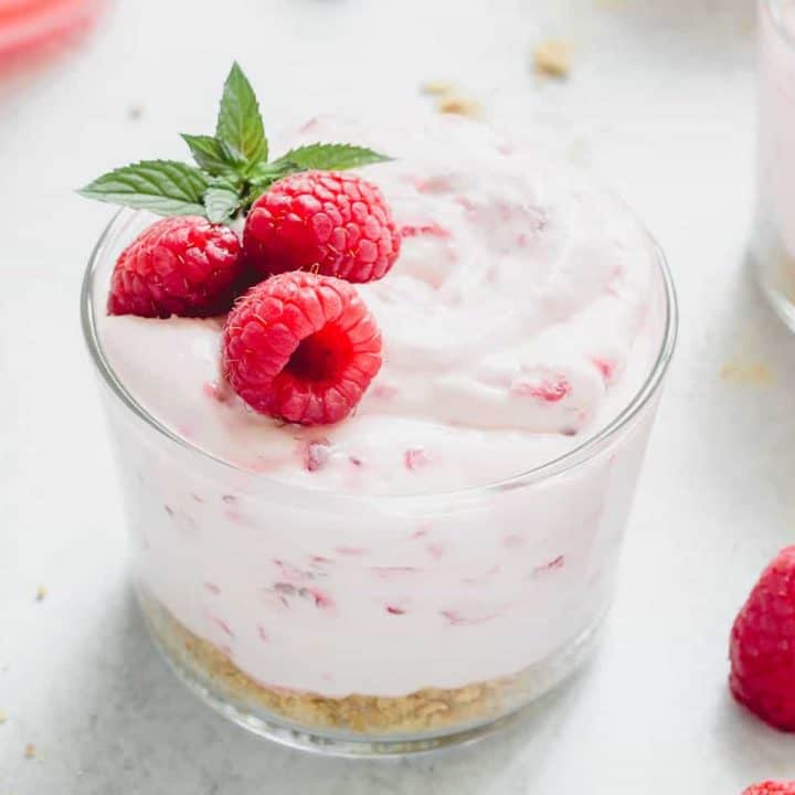 Close up of a glass dish of no-bake raspberry lemon cheesecake garnished with three raspberries and a sprig of mint