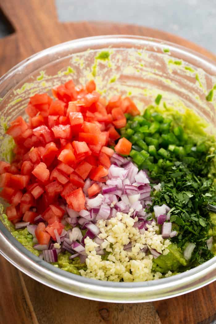 Tomato, onion, garlic, jalapeno and cilantro being added to mashed avocado in a glass mixing bowl