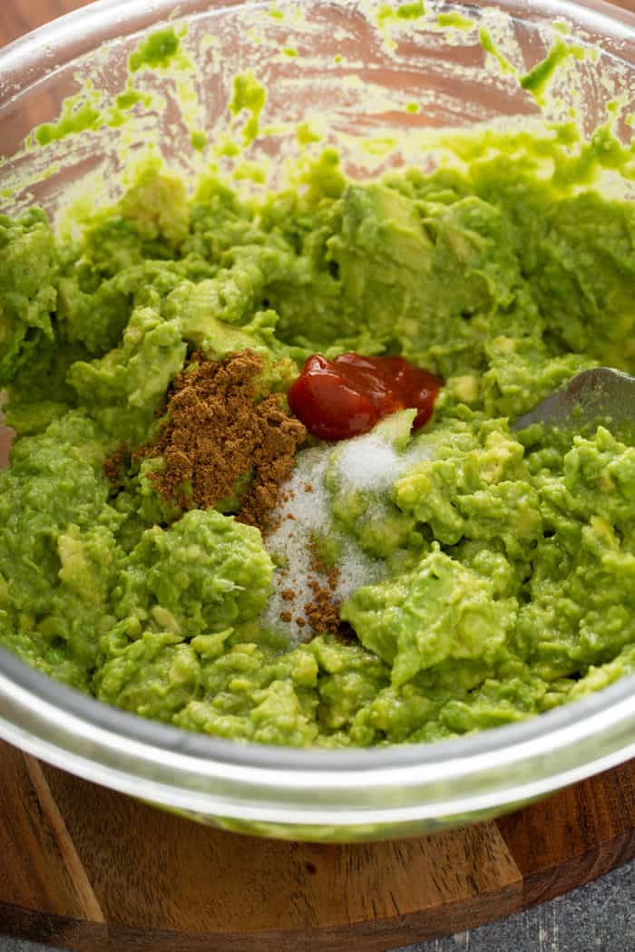 Salt, cumin, and sriracha being added to mashed avocado and lime in a glass mixing bowl